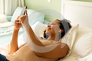 Happy biracial woman with earphones using smartphone and lying on bed in bedroom