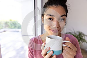 Happy biracial woman holding cup of coffee at sunny home