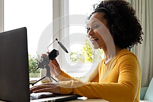 Happy biracial woman using headphones, laptop and microphone podcasting from home