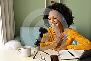 Happy biracial woman using headphones, tablet and microphone podcasting from home