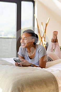 Happy biracial woman using smartphone on bed in bedroom