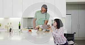 Happy biracial woman in wheelchair using tablet, preparing food with male partner in kitchen