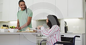 Happy biracial woman in wheelchair using tablet, preparing food with smiling male partner in kitchen