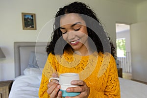 Happy biracial woman in yellow sweater holding cup of coffee at home