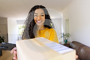 Happy biracial woman in yellow sweater holding package and using smartphone at home