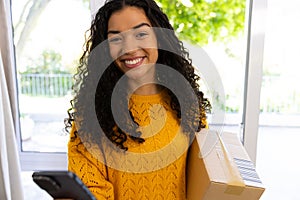 Happy biracial woman in yellow sweater holding package and using smartphone at home