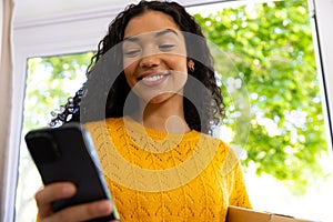 Happy biracial woman in yellow sweater holding package and using smartphone at home