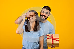 Happy birthday or anniversary. Happy arab man covering his wife's eyes, giving wrapped gift box, yellow background