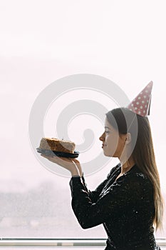 Happy birthday, beautiful young woman holding a cake looking at a candle, standing at the office window, light