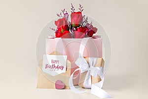 Happy birthday card and envelope with gift box and rose in paper bag on beige background