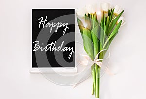 Happy Birthday card message sign on black chalkboard with tulip flowers on white background flat lay. Blackboard text