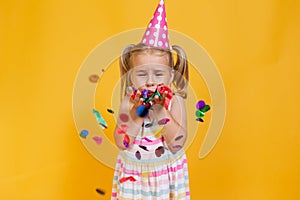 Happy birthday child girl in pink cup blowing confetti on colored yellow background. Celebration, childhood