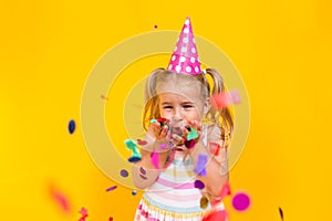 Happy birthday child girl in pink cup blowing confetti on colored yellow background. Celebration, childhood.