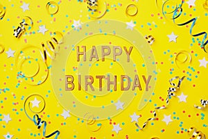 Happy birthday greeting text decorated with confetti and serpentine on yellow background top view. Flat lay style.