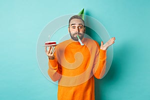 Happy birthday guy celebrating, wearing party hat, blowing wistle and holding bday cake, standing against white