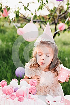 Happy birthday little girl making wish blowing candles on cake with pink decor in beautiful garden