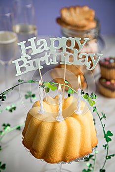 Happy birthday ring cake, cookies, biscuits, muffins and champagne  with clover decoration on marble table and lilac background