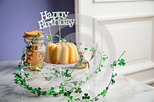 Happy birthday ring cake, cookies, biscuits, muffins and champagne  with clover decoration on marble table and lilac background