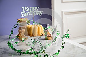 Happy birthday ring cake, cookies, biscuits, muffins and champagne  with clover decoration on marble table and lilac background