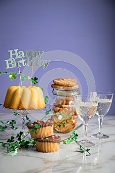 Happy birthday ring cake, cookies, biscuits, muffins and champagne  with clover decoration on marble table and lilac background