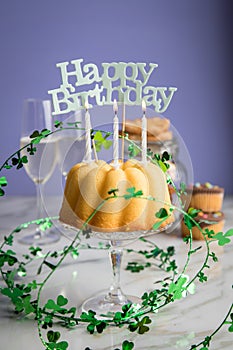 Happy birthday ring cake, cookies, biscuits, muffins and champagne  with clover decoration on marble table and lilac background