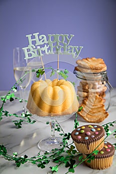 Happy birthday ring cake, cookies, biscuits, muffins and champagne  with clover decoration on marble table and lilac background