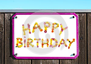 HAPPY BIRTHDAY sign made of yellow pink red and orange flowers collage, pink sign frame, wooden wall, turquoise sky