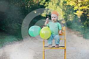 Happy birthday. Smiling little child boy in paper crown with balloons sitting on chair outdoor. Cute toddler celebrating birthday