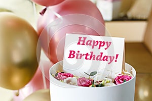 Happy Birthday text on gift card in flower box near festive pink and gold balloons. Beautiful bouquet of fresh flowers roses in