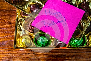 Happy birthday text on memo post reminder, near a golden box with chocolate pralines