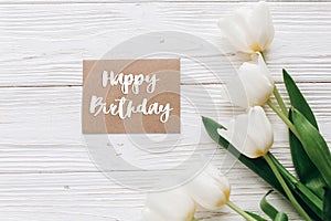 happy birthday text sign on stylish craft greeting card and tulips on white wooden rustic background. flat lay with flowers and g