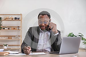 Happy black businessman talking on cellphone having phone conversation sitting at workplace working on laptop computer