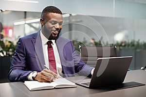 Happy black businessman using laptop and taking notes