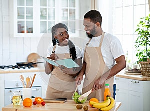 Happy Black Couple Checking Recipe In Cookbook While Cooking Together In Kitchen