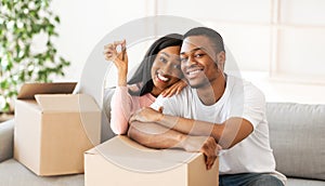 Happy black couple with house key and carton boxes looking at camera and smiling in their property, panorama photo