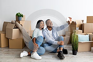 Happy black couple moving to their own home and taking selfie among cardboard boxes, free space