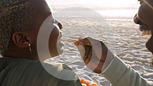 Happy black couple, sushi and beach picnic eating in romance on a sandy shore. African man feeding his wife seafood in