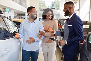 Happy Black Couple Talking To Salesman While Choosing Car In Dealership Center