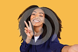 Happy black curly lady taking selfie and showing peace sign, against yellow background