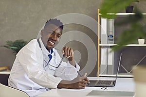 Young black doctor sitting at office desk with laptop, looking at camera and smiling