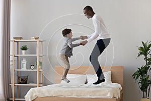 Happy black father and cute kid son jumping on bed