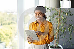 Happy black female entrepreneur using tablet standing near window at modern coworking office, copy space
