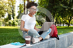 Happy Black Female Student Using Laptop Wearing Earbuds Sitting Outdoors