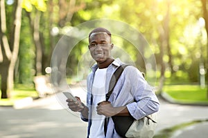 Happy black guy in casual outfit using mobile phone during his walk in city park