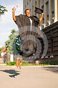 Happy black guy graduate from university with pleasure jumping up in black mantle and hat. Photo of student outdoors on
