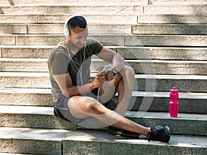 Happy black guy with headphones and mobile device relaxing on stairs and listening to music after training