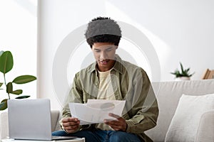 Happy black guy learning online from home, writing coursework, holding documents near laptop