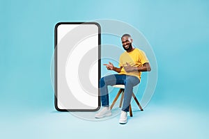 Happy black guy sitting on chair, pointing at giant smartphone with white screen, offering mockup for mobile app design