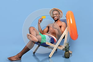 Happy black guy in swimwear showing credit card and gesturing thumb up while sitting in lounge chair, blue background