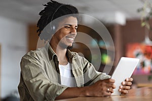 Happy Black Guy Wearing Wireless Headphones Using Digital Tablet At Cafe